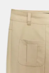 Pantalón de corte recto y largo tobillero, color beige. Presenta tiro medio, trabillas para cinturón, cierre frontal con cremallera y botón, y bolsillos tipo cargo de plastrón en el delantero.