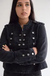 Chaqueta de estilo militar confeccionada en denim negro con efecto lavado. Presenta un diseño estructurado con cierre frontal de cremallera, cuello mao y múltiples botones metálicos decorativos dispuestos en filas horizontales. Incluye detalles de charreteras en los hombros y puños con botones.