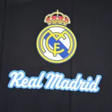 Remera de fútbol negra con cuello redondo y mangas cortas, con el escudo del Real Madrid estampado en el pecho y detalles en azul en los hombros.