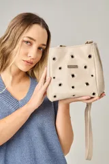 Bandolera de cuero color beige con diseño de ojales metálicos distribuidos por toda la superficie frontal y placa metálica con logo de la marca. Cuenta con correa larga ajustable.