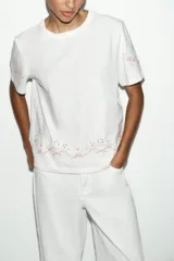 Camiseta de algodón de corte regular, color blanco, con cuello redondo y manga corta. Presenta detalles de bordados florales y ornamentales en color rosado en el ruedo y en las mangas.