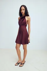 Vestido corto de tejido satinado color bordó, con cuello halter, espalda descubierta y detalle de frunces en la cintura.