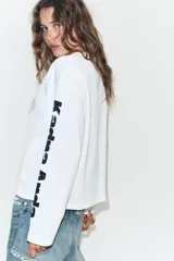 Camiseta blanca de corte oversize con cuello redondo y manga larga. Presenta una estampa gráfica central de una figura femenina, detalles estampados en la manga derecha y texto en la manga izquierda.
