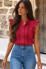 Blusa de diseño cuadrillé en color rojo y blanco, sin mangas, con volados en el cuello mao y en los hombros. Cuenta con alforzas verticales en el frente y cierre frontal de botones.