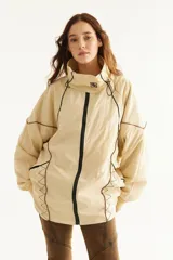 Campera rompevientos beige de nylon con diseño oversize, cuello alto, cierre frontal negro y detalles de costuras y ribetes en contraste.