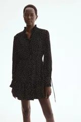 Vestido corto negro con estampado de lunares blancos, escote en V con volados, mangas largas abullonadas y cordón ajustable en la cintura.