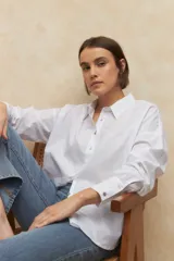 Camisa de corte clásico y holgado, confeccionada en tela blanca, con cuello camisero, cierre frontal de botones de nácar y mangas largas con puños abotonados.