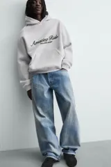 Sudadera con capucha y bolsillo frontal tipo canguro, confeccionada en tejido de algodón con interior perchado. Presenta diseño de corte boxy fit, mangas largas con puños y ruedo en rib, y texto bordado y estampado en el frente.
