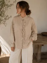 Camisa de color beige con cuello mao, cierre frontal de botones y detalle de volado vertical en el frente. Presenta mangas largas con puños abotonados y un corte relajado.