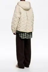 Chaquetón corto de corte línea A, confeccionado en tejido técnico acolchado color beige claro, con costuras en patrón geométrico. Posee cuello subido con capucha integrada, cierre frontal oculto con botones de presión, manga larga con puño elástico y vivos de ribete verde lima al contraste.