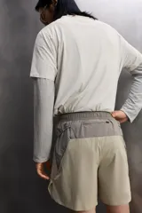 Pantalón corto de trail running color beige, confeccionado en tejidos técnicos ligeros y elásticos. Cuenta con forro interior, cintura elástica ajustable con cordón, bolsillos traseros laterales multifunción, bolsillo trasero central con cremallera, bajo con costura termofijada y aberturas laterales. Incluye logo estampado en la pernera.