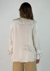 Blusa de satén color blanco con escote en V, cuello con volados y mangas largas con puños también decorados con volados.