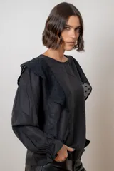 Blusa blanca de algodón con cuello redondo, mangas largas con puños elásticos y volados decorativos en los hombros.