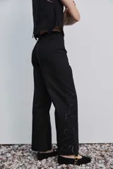 Pantalón negro de tiro alto y corte recto, confeccionado con detalles de bordados florales calados en la parte inferior de las piernas.