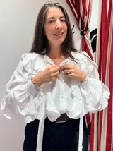 Camisa blanca de manga larga con volados frontales y lazos colgantes. Presenta mangas abullonadas con puños ajustados y cierre frontal con botones.