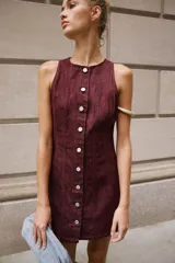 Vestido corto de denim color bordó, sin mangas, con cuello redondo, costuras pespunteadas a contratono y cierre frontal con fila de botones metálicos.