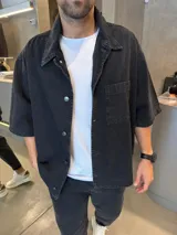 Camisa de jean de corte boxy y diseño holgado, color negro con efecto lavado. Presenta cuello clásico, cierre frontal con botones metálicos y un bolsillo frontal en el pecho.