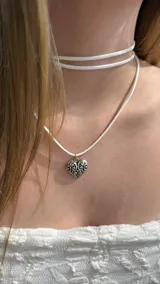 Gargantilla tipo choker de cordón blanco con dije de corazón metálico plateado con diseño de filigrana.