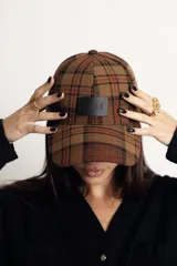 Gorra de béisbol con diseño de cuadros tartán en tonos marrones y rojizos, con parche frontal de cuero sintético negro con la marca grabada.