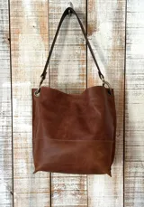 Cartera tipo bolso de hombro confeccionada en cuero marrón, con diseño minimalista, costuras visibles y herrajes metálicos en tono bronce.