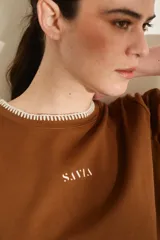 Remera marrón de manga corta y cuello redondo, con detalles de costuras decorativas en contraste color crudo en el cuello, los puños y el dobladillo inferior. Cuenta con el nombre de la marca impreso en el pecho.
