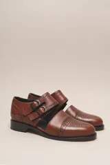 Zapato tipo monk strap con aberturas laterales, confeccionado en cuero vacuno metalizado color bronce. Presenta detalles de perforaciones estilo brogue en la puntera y el talón, y doble hebilla metálica en el empeine.