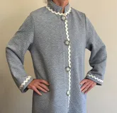 Bata de algodón capitoneado en color gris, con diseño de textura en zigzag. Presenta cuello mao, cierre frontal con botones y detalles de cinta decorativa en forma de ondas en cuello, puños y a lo largo de la abertura frontal.