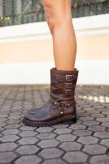Bota corta de cuero marrón oscuro, estilo motociclista, con caña media arrugada y tres hebillas ajustables en el lateral. Posee una tira decorativa con argolla metálica alrededor del tobillo y suela track gruesa.