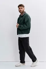 Cazadora de estilo urbano en color verde oscuro, con cuello de pana en tono marrón, cierre frontal con cremallera y puños elásticos. Presenta un diseño holgado con bordado pequeño en el pecho.