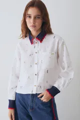 Camisa de lino blanca con estampado de motivos deportivos, cuello y puños tejidos a mano en crochet azul con borde rojo. Presenta cierre frontal con botones y un bolsillo en el pecho.