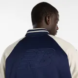 Campera tipo varsity New Balance, con cuerpo en azul marino y mangas en color crema. Presenta cierre frontal con botones a presión, cuello, puños y ruedo acanalados con rayas en contraste, y logo bordado en el pecho.