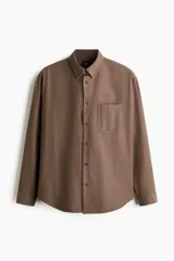 Camisa marrón de manga larga, confeccionada en sarga suave con cuello inglés, cierre frontal de botones, bolsillo en el pecho y bajo redondeado.