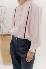 Camisa de algodón blanca con cuello a la base, cierre frontal con botones y mangas largas con puños delicados. Presenta volados verticales y detalles de cintas de terciopelo en contraste a lo largo del frente.