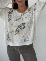 Buzo de tejido de punto abierto color beige, con diseño holgado, cuello redondo y mangas largas con puños acanalados. Presenta un estampado artístico que combina motivos de animal print y detalles brillantes en color dorado.