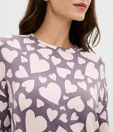 Conjunto de pijama compuesto por una blusa de manga larga con cuello redondo y un pantalón con cintura elástica. Ambas piezas presentan un diseño estampado de corazones en tonos lila y crema.