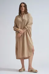 Camisa larga de lino color beige, de corte oversize, con mangas largas, cuello camisero y cierre frontal de botones. Presenta dobladillo curvo con aberturas laterales.