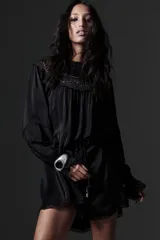 Vestido corto negro con cuello redondo, mangas largas abullonadas con puños elásticos y detalle de encaje y bordados con abalorios. Incluye lazo para ajustar en la cintura.