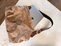Cartera tipo hobo o de hombro de cuero color camel con efecto gastado. Presenta costuras decorativas en contraste color celeste y un anillo metálico dorado en el frente. Incluye correa ajustable de cuero marrón oscuro con hebilla dorada.