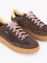 Zapatilla de cuero descarne color rosa, con suela de caucho color caramelo. Presenta cordones oscuros con un aplique de cristal decorativo y detalle de cuero liso en el talón.