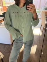 Blusa de manga larga color blanco con cuello redondo y cierre frontal de botones. Presenta detalles de bordado estilo punto smock en el pecho y textura de rayas verticales en la parte superior. Diseño holgado y estilo bohemio.