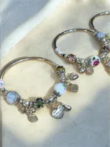 Pulsera rígida plateada con dijes colgantes y cuentas decorativas. Incluye cuentas de piedra veteada, cristales facetados en tono verde petróleo, una cuenta de cerámica blanca con diseño floral y múltiples dijes de corazón, uno de ellos con la frase 'you love you' grabada.