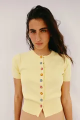 Chaqueta de punto color amarillo claro, con cuello redondo y manga corta. Presenta cierre frontal con botones de presión con diseño de flores de colores.