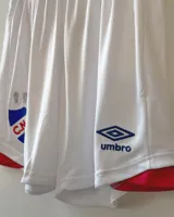 Short de fútbol Umbro del Club Nacional de Football, modelo Home 2025. Diseño en color azul con detalles en rojo en los laterales y el escudo del club junto al logo de la marca.