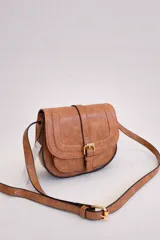 Bandolera de tamaño pequeño en color camel con acabado texturizado. Presenta un diseño de solapa frontal con detalle de hebilla decorativa y cierre magnético. Incluye una correa larga ajustable y herrajes metálicos en tono dorado.
