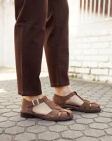 Sandalias de cuero gamuzado color marrón, tipo romanas, con tiras entrelazadas, talón cerrado y tira ajustable con hebilla metálica en el tobillo.