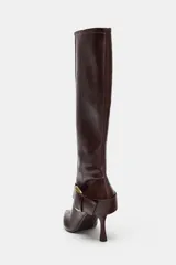 Bota de caña alta en color marrón oscuro, confeccionada en cuero con acabado liso. Presenta un diseño de punta fina, tacón alto fino y una tira decorativa en el tobillo con una hebilla metálica dorada. Incluye cierre lateral mediante cremallera.