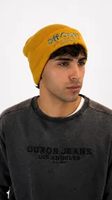 Gorro tipo beanie de punto color mostaza con bordado frontal en tonos azul y blanco.