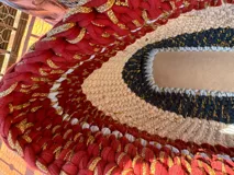 Bolso tejido a mano con hilo de trapillo, eco-cuero y textiles, con un diseño central de querubín coronado y alas, sobre un fondo de franjas rojas, blancas y azules. Posee asas cortas tejidas en color celeste claro.