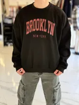 Buzo oversize negro de cuello redondo, con estampa frontal en tono rojizo con las inscripciones "BROOKLYN" y "NEW YORK".