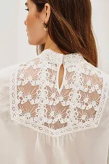 Camisa blanca de estilo romántico con cuello alto, confeccionada en algodón con detalles de encaje y bordados en el frente. Presenta mangas largas abullonadas con puños elásticos y terminaciones de encaje.